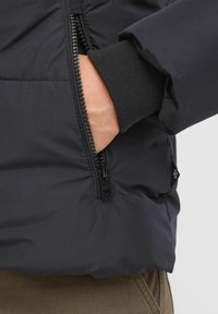 Doudoune noire avec un extérieur lisse, dotée d'une poche latérale zippée et de poignets côtelés. Le tissu semble léger et isolant.