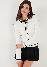 Weißer Strickcardigan mit schwarzem Besatz, Knopfverschluss und einer Schleife am Hals. Kombiniert mit einem gefalteten schwarzen Rock.