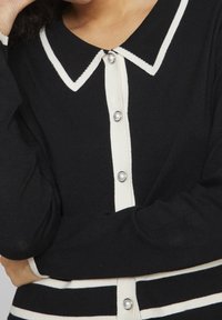 Svart stickad cardigan med vit krage, knappslå och horisontella vita ränder vid fållen och ärmarna. Det går att se pärl-liknande knappdetaljer.