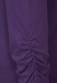 Lila långärmad topp med samlade detaljer vid ärmsluten, tillverkad av mjukt bomullsmaterial med en slät yta.