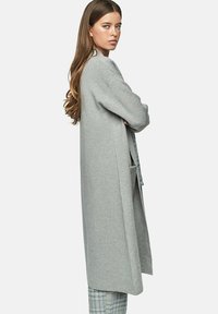 Long cardigan gris en tissu doux, avec une coupe ample, des manches larges et des poches latérales. Stylé sur un pantalon à carreaux.