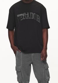 Mies, joka on pukeutunut mustaan ylisuureen T-paitaan, jossa on "PEGADOR"-teksti ja pieni motivoiva lainaus, yhdistettynä harmaisiin cargo-housuihin.