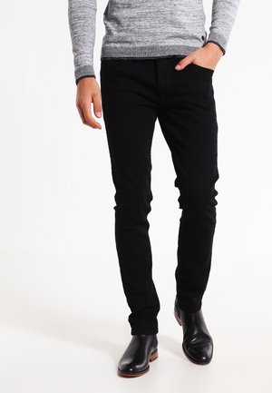 Hombre vestido con pantalones negros ajustados, suéter gris de manga larga, botas de cuero negras, con una mano en el bolsillo, sobre un fondo blanco.