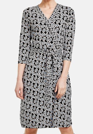 Femme portant une robe portefeuille mi-longue à manches trois-quarts, motif géométrique noir et blanc, ceinture nouée à la taille, debout devant un fond uni.