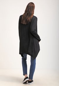 Černý dlouhý cardigan s volným zadním dílem, vyrobený z měkké látky; spárovaný se světle modrými džíny a černými teniskami.