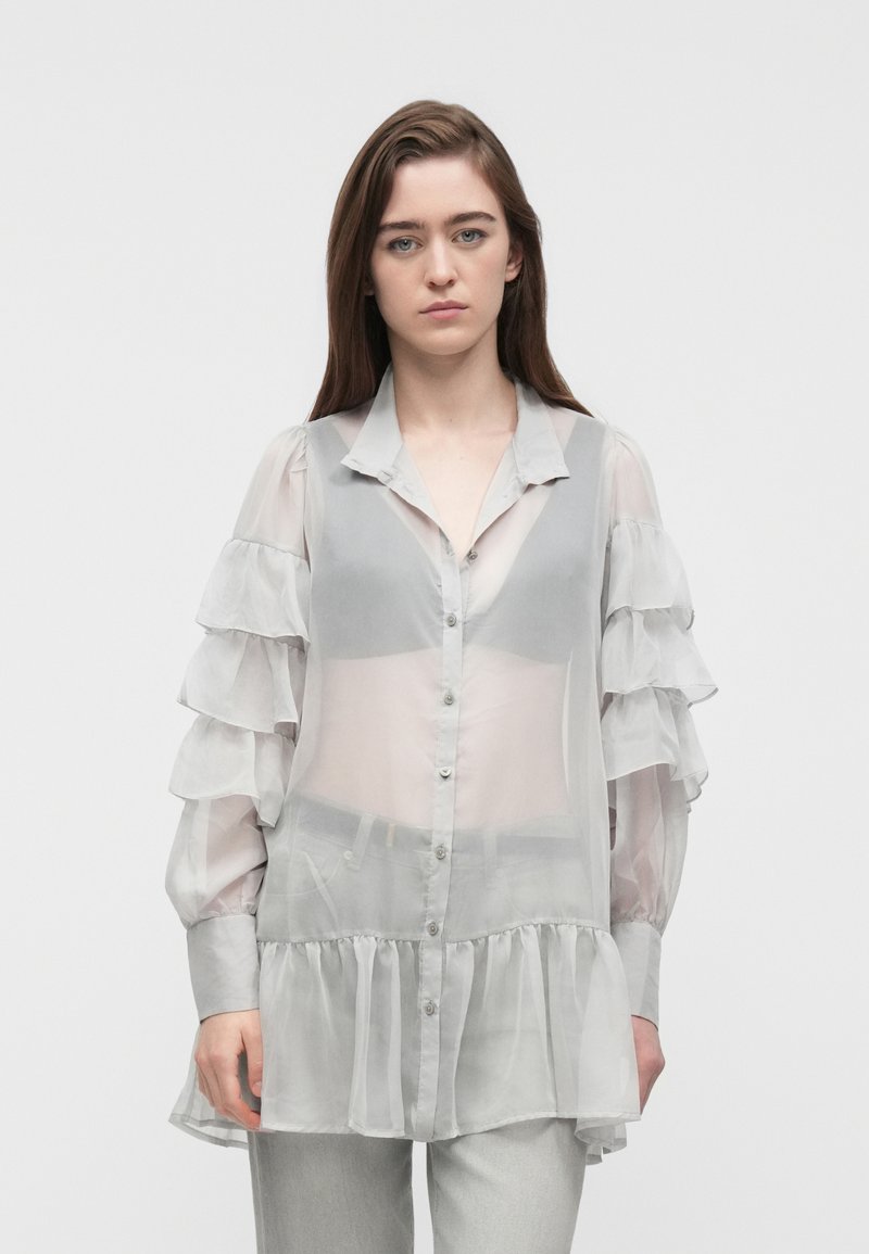 Νεαρή γυναίκα που φοράει μια διαφανή ανοιχτογκρίζα μπλούζα με κουμπιά και μανίκια με στρώσεις βολάν, καθώς και ανοιχτογκρίζο παντελόνι, στέκεται μπροστά σε απλό φόντο.