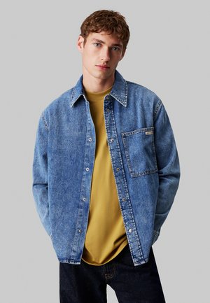 Giacca di jeans azzurro chiaro, con chiusura a bottoni, colletto aperto e taschino sul petto, indossata sopra una t-shirt gialla senape.