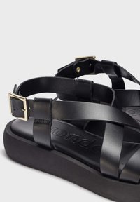 Sandales en cuir noir avec des brides croisées, fermeture à boucle en métal et une semelle épaisse et texturée noire présentant un logo en relief.