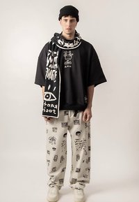 Čierne nadrozmerné tričko s grafickým dizajnom, biely šál s vzorom, béžové nohavice pokryté čiernym textom a obrázkami a biele tenisky.