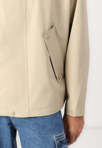 Beige lett jakke med en skjult lomme og knappelukking. Glatt tekstur, avslappet passform og lange ermer. Brukt over denim cargo-bukser.