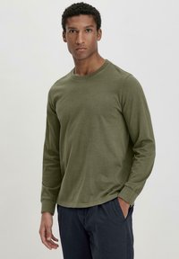 Olivgrünes Langarm-T-Shirt aus weichem Baumwollmaterial mit Rundhalsausschnitt und gerippten Bündchen, kombiniert mit dunkelblauen Hosen.