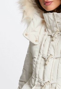 Giacca imbottita bianca con cappuccio foderato in pelliccia, dettagli in metallo dorato e chiusure a cordoncino. La texture è liscia con dettagli trapuntati.