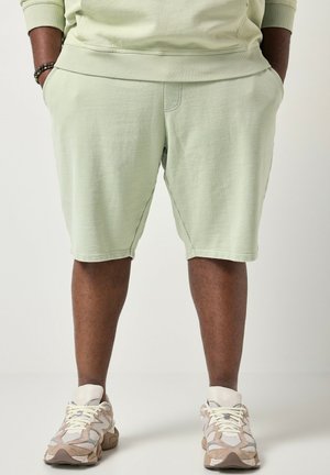 Person iført lysegrønne shorts og beige sportssko med hænderne i lommerne, stående mod en ensfarvet hvid baggrund.