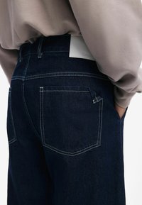 Dark blue jeans featuring a classic design with two back pockets, white leather tag, and contrasting stitching along the seams.