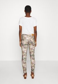 Getailleerde broeken met een bloemen- en dierenprint in groen, bruin en crème; gecombineerd met een witte shirt met korte mouwen en bruine sandalen met hak.