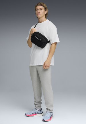 Ung mand iført hvid oversized t-shirt, grå joggingbukser, lyseblå sneakers, sort crossbody-taske og sort pandebånd, stående mod en grå baggrund.