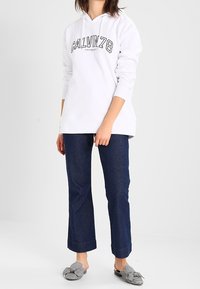 Bíla mikina s nápisem "CALVIN", modré džíny s ohrnutým lemem a šedé slip-on boty s vzorem. Jednoduchý, neformální outfit.