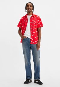 Hombre con cabello trenzado que lleva una camisa de botón roja de manga corta con patrones de hormigas blancas, una camiseta blanca, jeans azules y zapatillas negras.