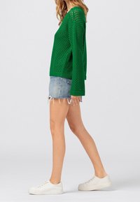 Kvinna som går i en grön perforerad tröja, trasiga denimshorts och vita sneakers mot en ljus enfärgad bakgrund.