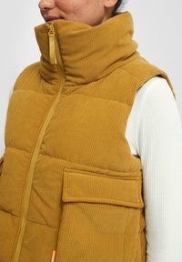 Person, der eine senfgelbe Cordweste mit hoher Kragen und einer Fronttasche über einem weißen gerippten Langarmshirt trägt.