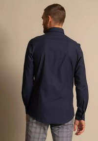 Mees kannab tumeda meresinist pika varrukaga särki ja halli ruudulist püksid, seisab kaameraga seljaga vastu beeži tausta.