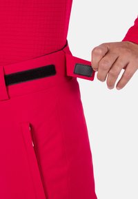 Helle pinke Hosen mit strukturiertem Design und einem Klettverschluss am Bund, mit einer seitlichen Reißverschlusstasche sichtbar.