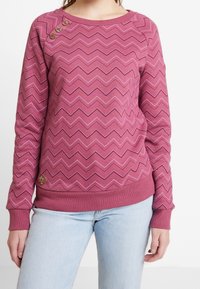 Mujer vistiendo un suéter de patrón en zigzag rosa con tres botones decorativos cerca del hombro izquierdo y jeans azul claros.