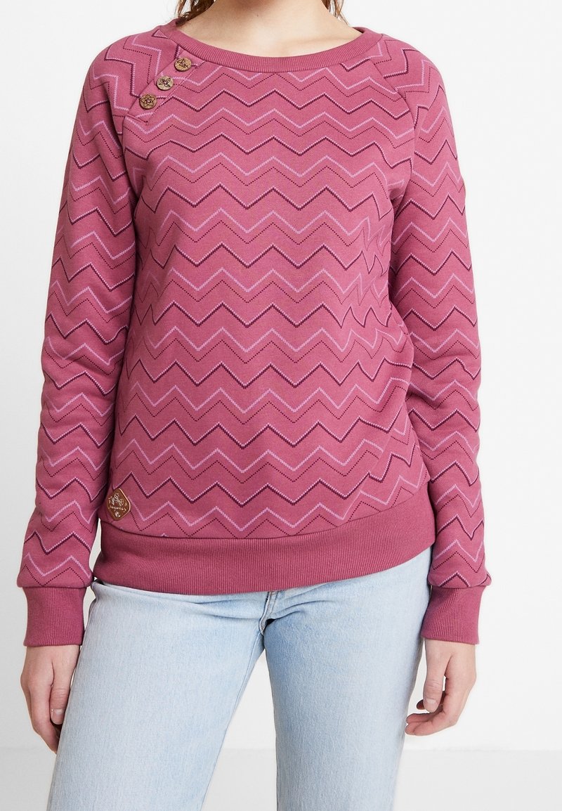 Mujer vistiendo un suéter de patrón en zigzag rosa con tres botones decorativos cerca del hombro izquierdo y jeans azul claros.