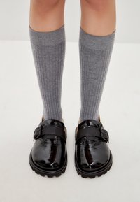 Zapatos de charol negro con hebilla, parte superior texturizada y suelas gruesas, combinados con calcetines de canalé gris hasta la rodilla.
