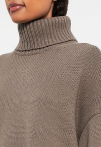 Femme portant un pull col roulé en tricot côtelé marron, mettant en valeur le décolleté et la partie supérieure de la poitrine.