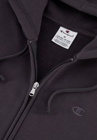 Tmavosivá mikina na zips vyrobená z mäkkej látky. Obsahuje predný zips, logo na hrudi a kapucňu na šnúrku. Viditeľný štítok na starostlivosť.