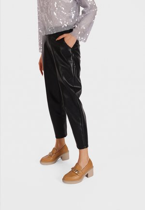 Pantaloni tapered in finta pelle nera, un maglione argentato con paillettes e mocassini con tacco color marrone chiaro e una catena dorata come accentuazione.