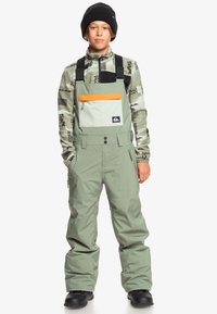 Groene sneeuwoverall met verstelbare banden, ritszakken en een oranje accentzak. Gestructureerde zwarte en groene patroononderlaag eronder.