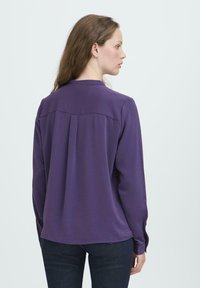 Donna con lunghi capelli castani che indossa una blusa viola a maniche lunghe e jeans scuri, rivolta di lato su uno sfondo chiaro e semplice.