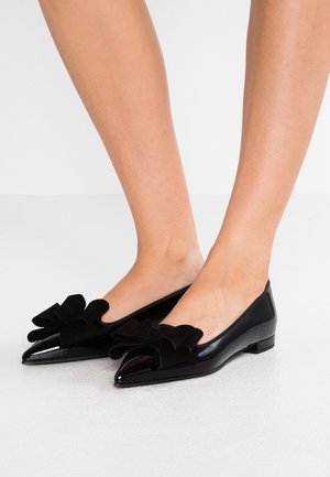 Chaussures plates en cuir verni noir à bout pointu portées aux pieds, ornées de grands nœuds en tissu noir sur la partie supérieure avant de chaque chaussure.