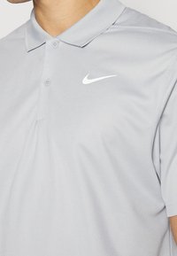 Světle šedé polo tričko Nike má klasický límec, dvě knoflíkové légie a bílé logo na levé straně. Hladká, na vlhkost odvádějící tkanina.