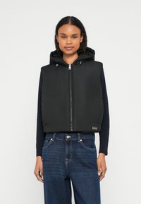 Μαύρο μπουφάν τύπου γιλέκο με κουκούλα, κλείσιμο με φερμουάρ και ρυθμιζόμενους κορδόνια, φορεμένο πάνω από ένα σκούρο μπλουζάκι με μακριά μανίκια και συνδυασμένο με μπλε τζιν.