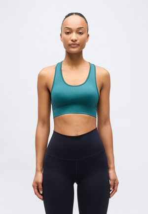 Femme debout, face à l'avant, portant un soutien-gorge de sport bleu sarcelle et un legging noir taille haute, devant un fond clair uni.