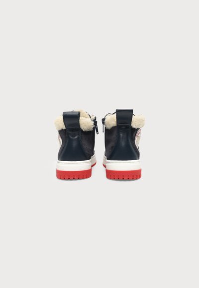 Paire de bottes d'hiver noires avec semelles rouges et doublure en fourrure crème, montrée de dos sur un fond clair uni.