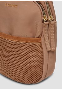 Zaino in tessuto beige con pannello in rete, cerniere dorate e accenti marroni. Presenta una superficie testurizzata e un fondo arrotondato per un design compatto.