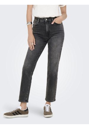 Personne portant un jean noir délavé taille haute et des baskets marron avec des rayures noires, debout avec une main dans la poche et l'autre reposant sur la cuisse.