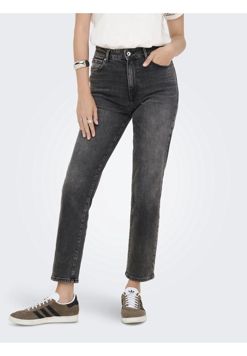 Personne portant un jean noir délavé taille haute et des baskets marron avec des rayures noires, debout avec une main dans la poche et l'autre reposant sur la cuisse.