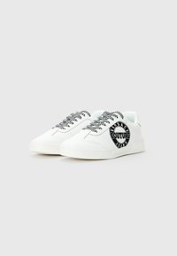 Biele nízkopriemerné tenisky s čierno-bielymi vzorovanými šnúrkami a čiernym kruhovým logom s nápisom "Versace Jeans Couture" na boku.