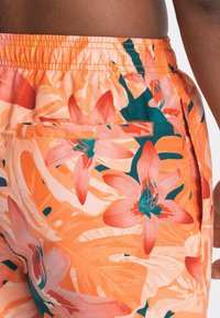 Gros plan sur une personne portant un short de bain orange avec des motifs floraux roses et turquoise et une ceinture élastique.
