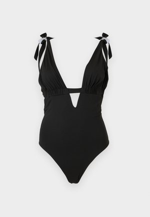 Fekete egyrészes fürdőruha mély V-nyakkivágással, mell alatt kivágással és vállpántokkal, egyszerű világos háttér előtt.