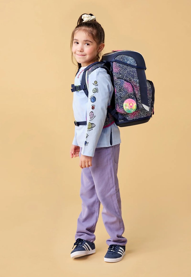 Sac à dos avec base marine, motif galaxie coloré, accents réfléchissants, sangles ajustables et poche avant zippée. Enfant portant un t-shirt à manches longues et un pantalon violet.
