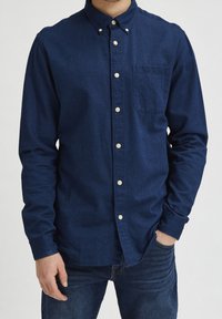 Tmavě modrá džínová košile na knoflíky s náprsní kapsou, bílými knoflíky a klasickým límečkem.