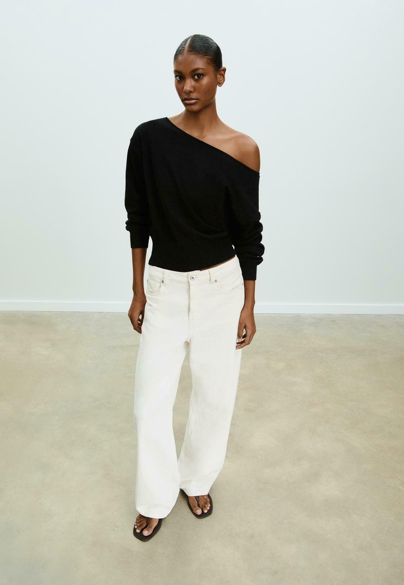 Maglione nero off-shoulder a maniche lunghe, abbinato a jeans bianchi larghi e infradito neri. Tessuto morbido, design informale.