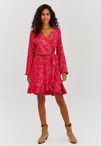 Červené dlouhé rukávy, zavinovací šaty s volánkovým lemem a texturovaným vzorem. Obsahují zavázání v pase. Model nosí béžové kotníkové boty.