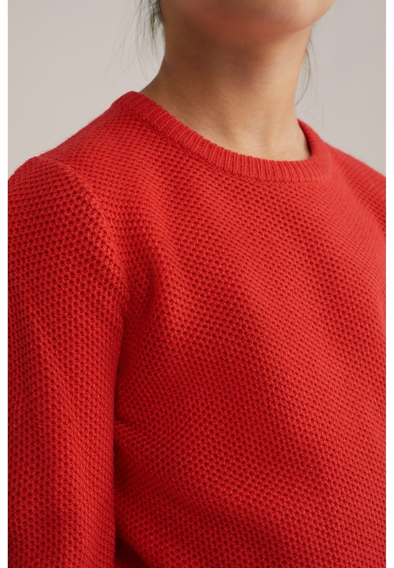 Gros plan d'une personne portant un pull tricoté rouge texturé avec un col rond sur un fond neutre.